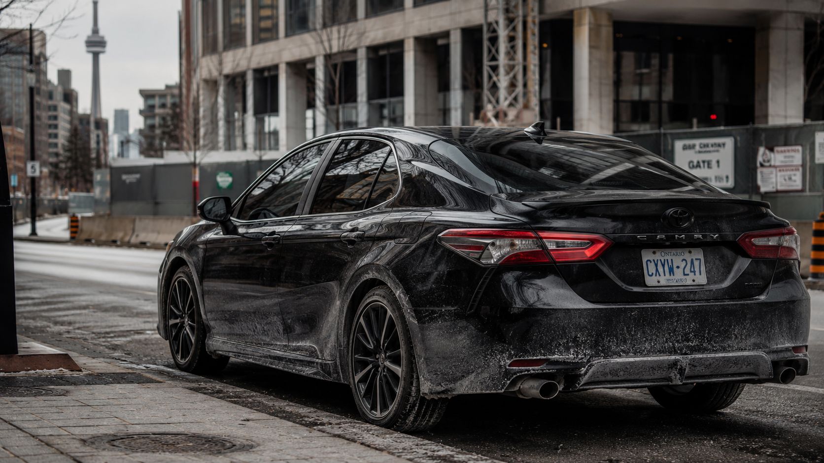 A black Toyota Camry parked on a snowy city street in Toronto with the CN Tower in view.