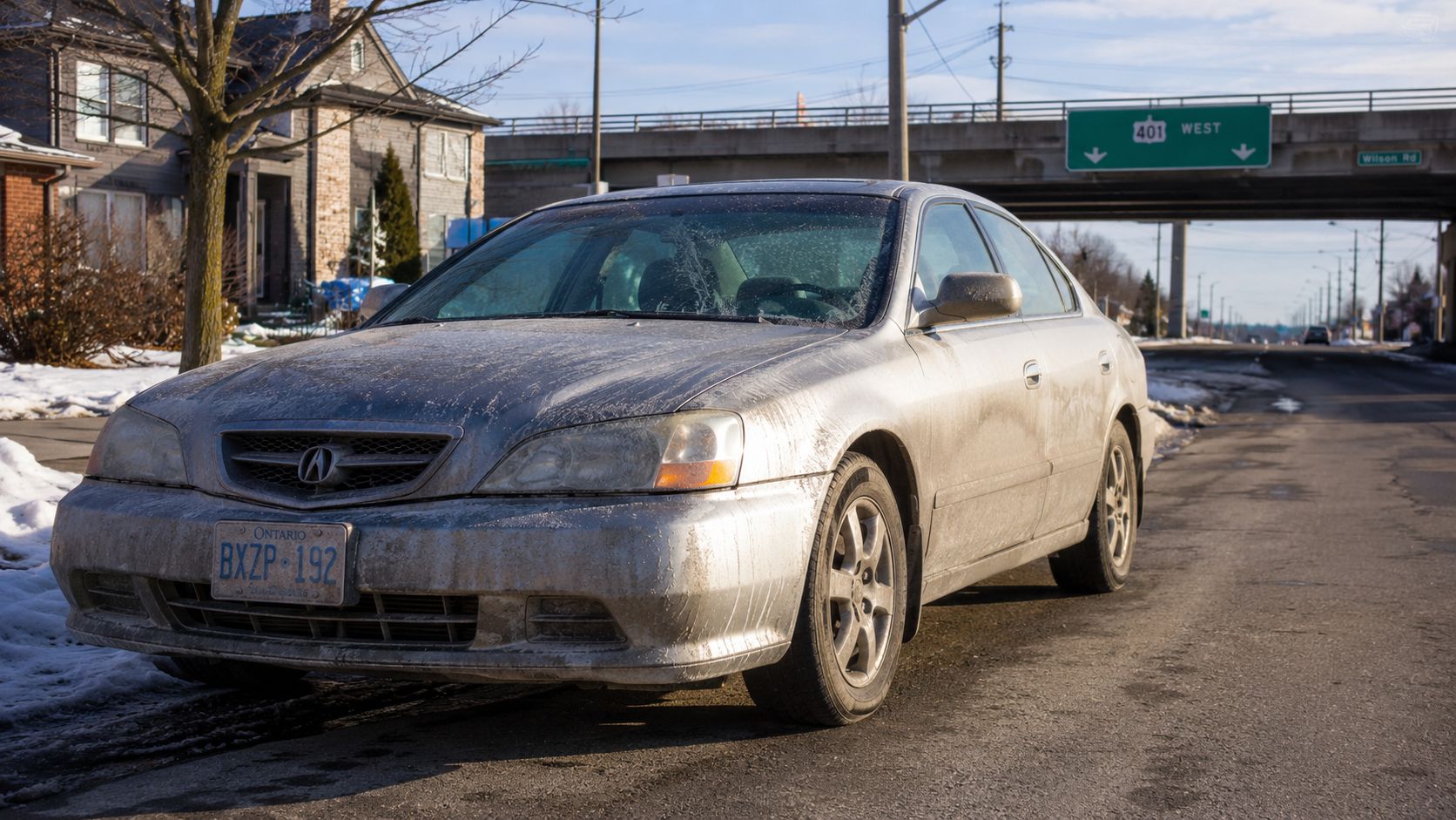 A dirty silver Acura sedan parked on the side of a road near a highway overpass in Ontario.