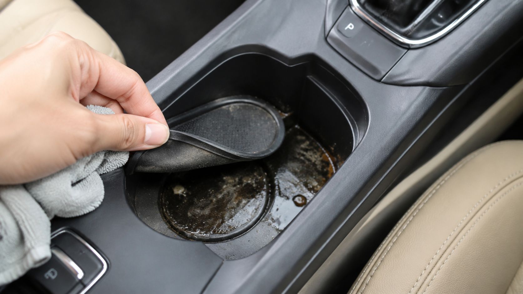 A person cleaning a dirty, sticky car cup holder with a grey microfiber cloth in the interior.