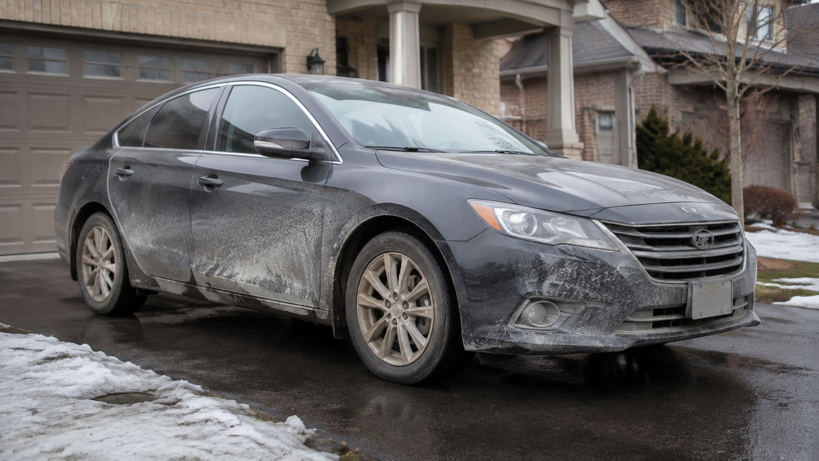 A dirty black sedan covered in road salt parked on a wet driveway during wintertime.