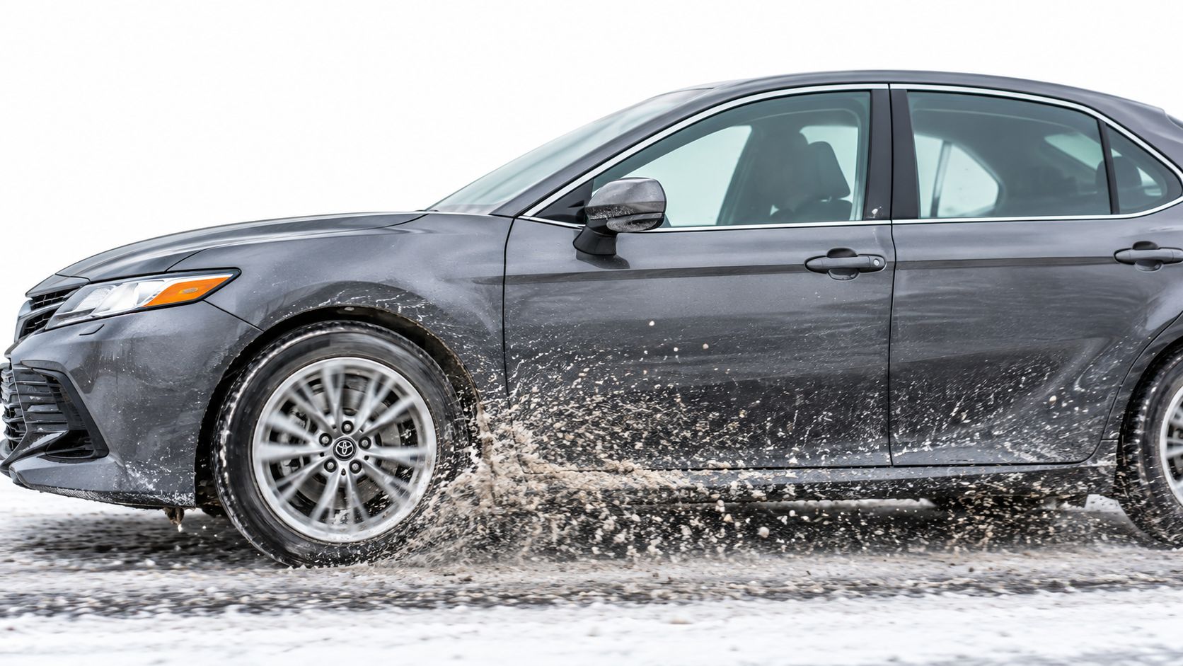 A gray Toyota sedan driving through slush on a winter road, splashing icy water from its tires.