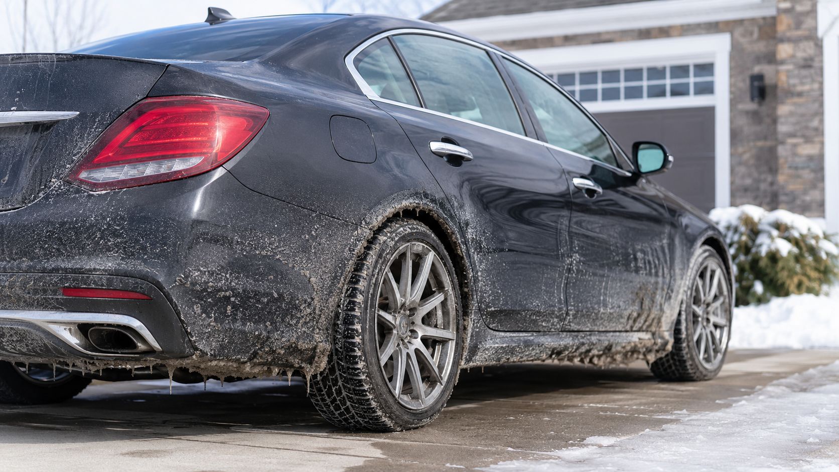 A dirty black Mercedes-Benz car parked in a snowy driveway with ice and road salt on the undercarriage.