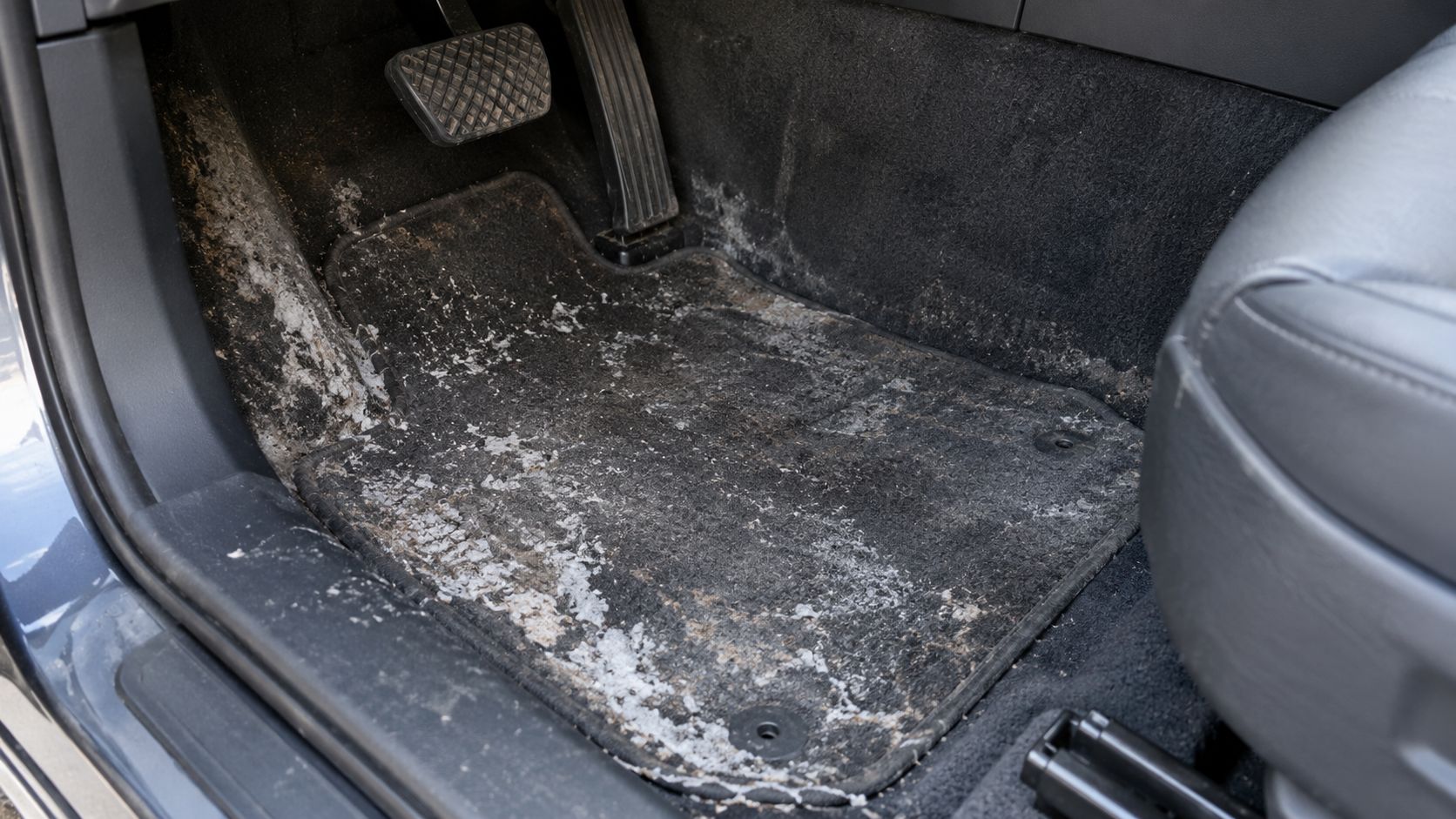 A dirty car floor mat covered in salt and dirt residue in the driver side footwell.