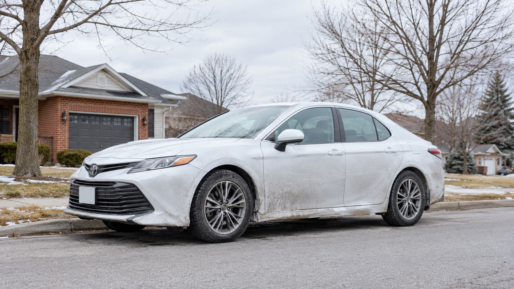 A dirty white Toyota Camry sedan parked on a residential street in a neighborhood during winter.